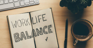 An open notebook with "Work Life Balance" written on it. A pen rests beside the notebook, and a cup of coffee is nearby on a desk. A computer keyboard and a potted plant are partially visible.