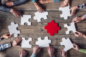 A group of hands, belonging to diverse individuals, is assembling white puzzle pieces around a single red piece on a wooden table. The focus is on teamwork and collaboration, with the red piece serving as the central element.