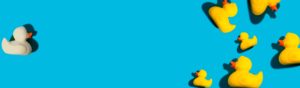 A group of yellow rubber ducks with red beaks is gathered in the bottom right corner of a bright blue background. A single white rubber duck with an orange beak is isolated on the left side, casting a shadow.