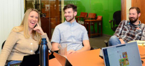 Three people sitting at a table in an office, smiling and engaged in conversation. Laptops and a water bottle are on the table. The background has a green wall with wooden chairs.