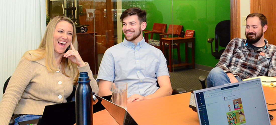 Three people sitting at a table in an office, smiling and engaged in conversation. Laptops and a water bottle are on the table. The background has a green wall with wooden chairs.