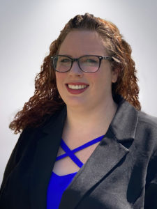A woman with wavy brown hair wearing glasses and a black blazer over a blue top smiles at the camera. She stands against a plain light background.