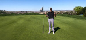 A man stands next to a flagstick on a golf green, holding up both index fingers. He is wearing a cap, sunglasses, a black shirt, gray pants, and white shoes. The course stretches into the distance under a clear blue sky.