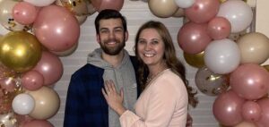 A smiling couple poses in front of a decorative balloon arch. The balloons are a mix of pink, gold, and white colors. The woman is wearing a light pink blouse and has her hand on the man's chest. The man is wearing a blue and black checkered shirt over a hoodie.