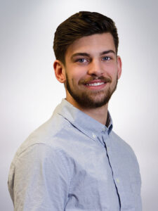 A man with a beard and short hair smiles, wearing a light gray button-up shirt against a neutral background.