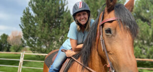 A woman in a blue shirt and jeans is smiling while sitting on a brown horse. She is wearing a black helmet, and there are green trees in the background. The scene appears to be outdoors in a fenced area.