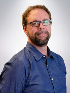 A man with a beard and glasses is standing against a light background. He is wearing a denim shirt and looking slightly to the side with a neutral expression.