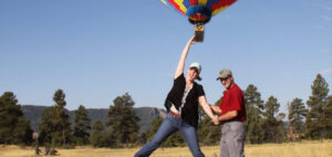 A woman playfully hangs onto a rope from a colorful, grounded hot air balloon. She's smiling and holding hands with a man in a red shirt and cap. The background features a field and trees under a clear blue sky.
