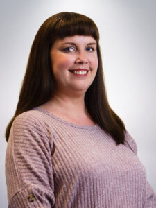 Smiling person with long brown hair and bangs, wearing a light purple ribbed sweater, against a soft gray background.