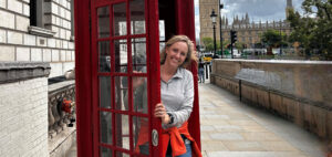 A smiling woman stands in the doorway of a classic red British phone booth on a sidewalk, with the UK Parliament buildings visible in the background.