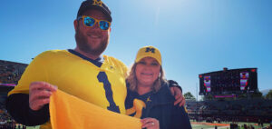 A man and a woman stand in a stadium wearing matching yellow and blue sports apparel, including hats with an "M" logo. They hold up a yellow flag or cloth and smile at the camera, with a scoreboard visible in the background.