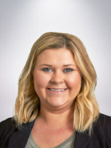 A smiling person with shoulder-length blonde hair is wearing a dark blazer over a gray top. The background is a light gradient.