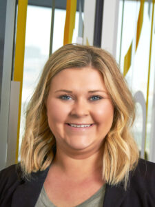 Smiling person with shoulder-length blonde hair wearing a black blazer and gray top, standing indoors with a glass wall featuring yellow accents in the background.