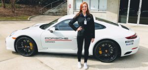 A person stands next to a white Porsche 911 GTS with "Porsche Experience" written on the side. The car has black wheels with yellow brake calipers. They are outdoors near a modern building.