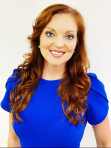 Woman with long red hair wearing a bright blue dress, smiling against a plain white background.