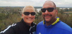 A couple wearing sunglasses smiles for a selfie outdoors. The man is in a blue jacket, and the woman in black. Behind them, trees and a river are visible under a cloudy sky.