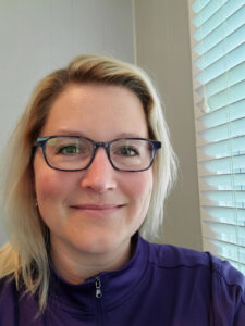 A person with blonde hair and glasses is smiling at the camera. They are wearing a purple top and are indoors near a window with blinds.