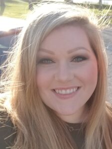 A woman with long blonde hair smiles at the camera in an outdoor setting with sunlight highlighting her hair. She is wearing a dark top, and the background is slightly blurred.