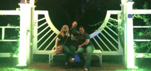 Three people sitting on a large white bench under a lit archway at night. Two individuals are seated on the bench, and one person is standing behind them, smiling. The scene is surrounded by foliage.