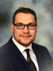 A man with glasses, wearing a navy suit, white shirt, and dark patterned tie, poses against a blurred gray backdrop. He has short brown hair and a light beard, and is smiling gently at the camera.