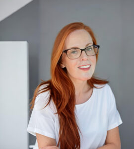 A woman with long red hair and glasses smiles while standing in front of a gray and white background. She is wearing a white shirt.
