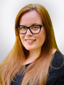 A smiling person with long, straight red hair and glasses, wearing a black top. The background is a soft gradient, enhancing the subject's features.