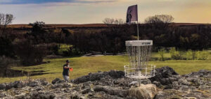 A person in a grey shirt stands on a rocky terrain playing disc golf. They are preparing to throw a disc towards a metal basket with chains and a flag on top. The background features trees and an overcast sky.