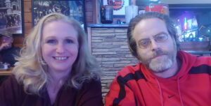A woman with long blonde hair and a man with glasses and a beard sit together at a restaurant. She is smiling, while he has a neutral expression. The background features stone decor and dim lighting.