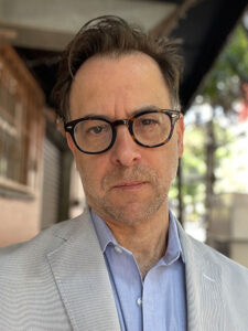 A man wearing glasses and a light gray suit jacket over a blue shirt stands outdoors. Behind him, the background features an out-of-focus urban setting with trees and a building.