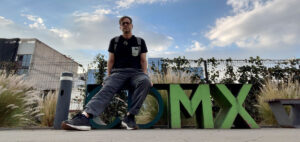 A person sitting on large, colorful letters spelling "CDMX" with a backdrop of plants and a partly cloudy sky. The scene is urban, with buildings partially visible in the background.