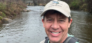 A man wearing a cap and backpack smiles while standing in shallow water of a flowing river. The background shows a bridge and trees lining the riverbank.