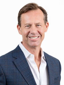 A smiling man in a blue checkered suit and white shirt stands against a plain white background.