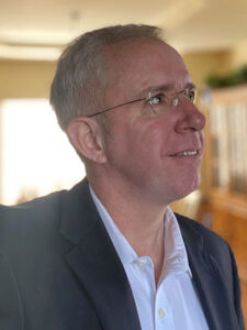 A man with short gray hair and glasses is looking thoughtfully upwards. He is wearing a dark suit jacket over a white shirt. The background is softly lit, suggesting an indoor environment.