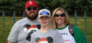 A family of three poses happily outdoors. The father wears a red cap and sunglasses, the son sports a baseball cap and reflective sunglasses, and the mother has long blonde hair and sunglasses. All are wearing festive shirts with holiday designs.