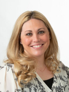 A woman with long blonde hair smiles at the camera. She is wearing a light-colored blouse with a floral pattern and delicate jewelry. The background is plain white.