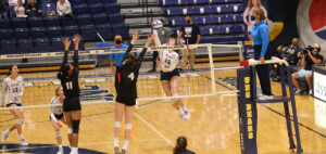 Two volleyball teams compete in a gym. A player in a white jersey jumps to spike the ball over the net, while two players in black jerseys attempt to block. A referee observes from an elevated position. The court reads "UNC Bears.