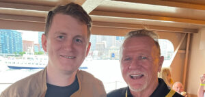 Two men are smiling on a boat with a city skyline and water in the background. The man on the left has short hair and is wearing a tan jacket. The man on the right has a goatee and is wearing a black shirt. A ferry and buildings are visible in the distance.