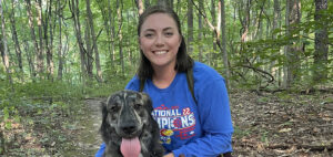 A woman in a blue "National Champions" shirt kneels beside a happy, panting dog on a forest trail. The background is filled with lush green trees and foliage.