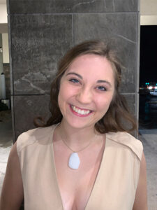 A smiling woman with long brown hair stands in front of a tiled wall. She is wearing a beige top and a necklace with a large, white pendant. The background appears to be outside at night with some lights visible.