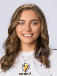 A person with long wavy hair smiles at the camera, wearing a white shirt with a softball logo.