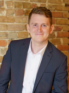 A person with short hair is smiling and wearing a white shirt with a dark blazer. They are standing in front of a brick wall.