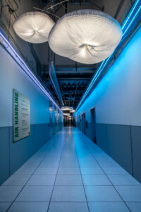 A futuristic corridor with tiled floors and glowing blue lights along the walls. The ceiling features large, circular light fixtures made of a net-like material. A sign labeled "Air Handling" is visible on the left wall.