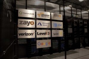 Image of a data center interior with various company logos on a metal grid wall, including CenturyLink, Amazon Web Services, Zayo, Microsoft Azure, AT&T, Verizon, Level 3, Sprint, and Google. Networking equipment is visible in the background.