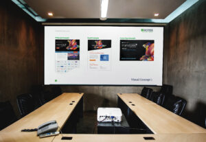 A modern conference room with a large screen displaying a presentation titled "Visual Concept 1," featuring examples of webpage, email, and cover page designs. A projector is placed on a long, polished wooden table surrounded by black chairs.