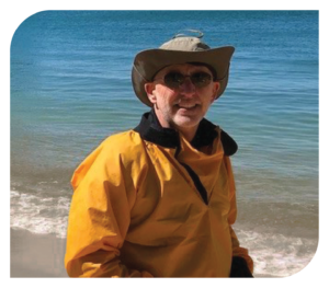 A man wearing a yellow jacket, a wide-brimmed hat, and sunglasses stands on a beach with the ocean in the background.