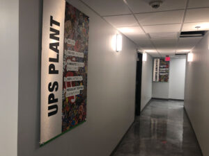A hallway with grey walls and polished floors. Two colorful vertical posters are mounted on the walls. The nearest one reads "UPS PLANT" and has various phrases listed. Ceiling lights illuminate the corridor. Exit signs are visible in the distance.