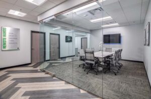 A modern office meeting room with glass walls. Inside, a rectangular table surrounded by several chairs is visible. A large screen is mounted on one wall. The floor has a patterned design, and fluorescent lights illuminate the room.