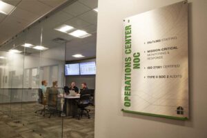 A glass-walled operations center shows four people seated around a table with computer screens. A wall sign reads "Operations Center NOC," highlighting features like 24/7 staffing, ISO 27001 certification, and SOC 2 audits.
