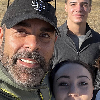 A group selfie with four people outdoors. The person in front is wearing a dark cap and jacket. They all appear to be smiling or content. A field is visible in the background under natural light.