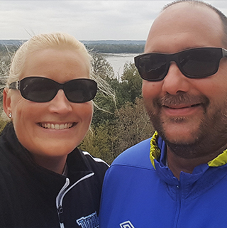 A woman and a man wearing sunglasses smile for a selfie outdoors. They are dressed in casual jackets and standing in front of a scenic view with trees and a body of water in the background.